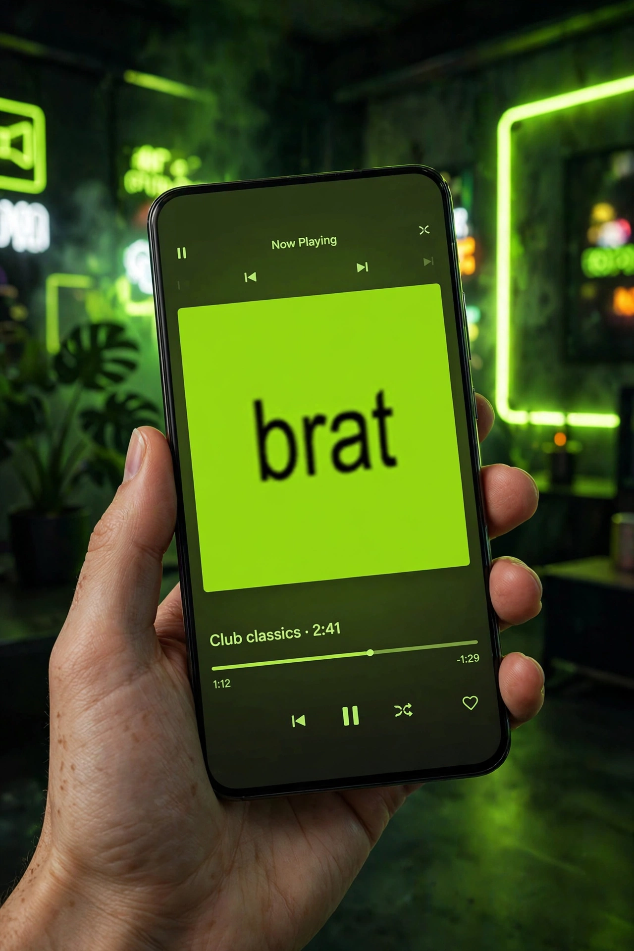 Hand holding a smartphone with a music player screen showing a lime-green brat-style album cover that reads 'brat', in a neon-lit room